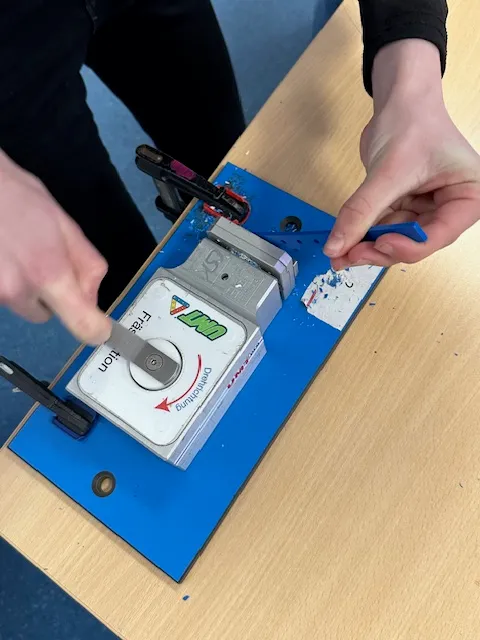 Hände montieren an einem blauen Holzbrett ein graues Gerät mit einem roten Aufkleber und Pfeil, befestigt mit zwei Schraubzwingen.