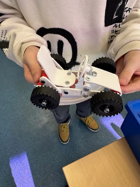 Ein Kind hält einen selbstgebauten Strandbuggy aus weißen und grauen Bauteilen mit großen schwarzen Reifen in der Hand.