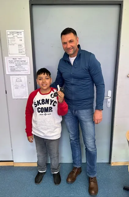 Ein Mann und ein Junge stehen vor einer Tür und halten ein kleines ferngesteuertes Auto in der Hand. Im Hintergrund ist ein Schild mit Informationen zu Räumen und Technik zu sehen.