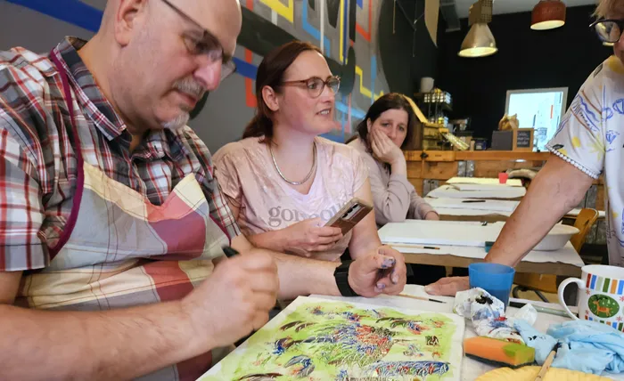 Ein Mann und eine Frau sitzen an einem Tisch und malen mit Wasserfarben. Im Hintergrund sind weitere Personen und Kunstwerke zu sehen.