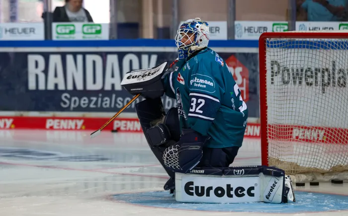 Eishockey-Torwart Hendrik Hane kniet in teal-farbener Ausrüstung vor dem Tor auf dem Eis.