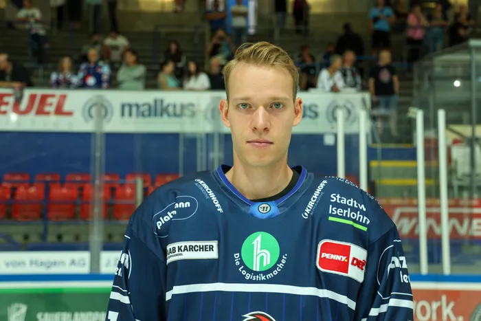 Maximilian Eisenmenger in seinem blauen Eishockeytrikot vor einer unscharfen Zuschauertribüne in einer Eishalle.