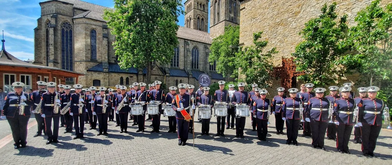 Eine große Blaskapelle in Uniformen steht auf einem Platz vor einer gotischen Kirche. Die Musiker spielen verschiedene Instrumente, darunter Trompeten, Posaunen und Trommeln.