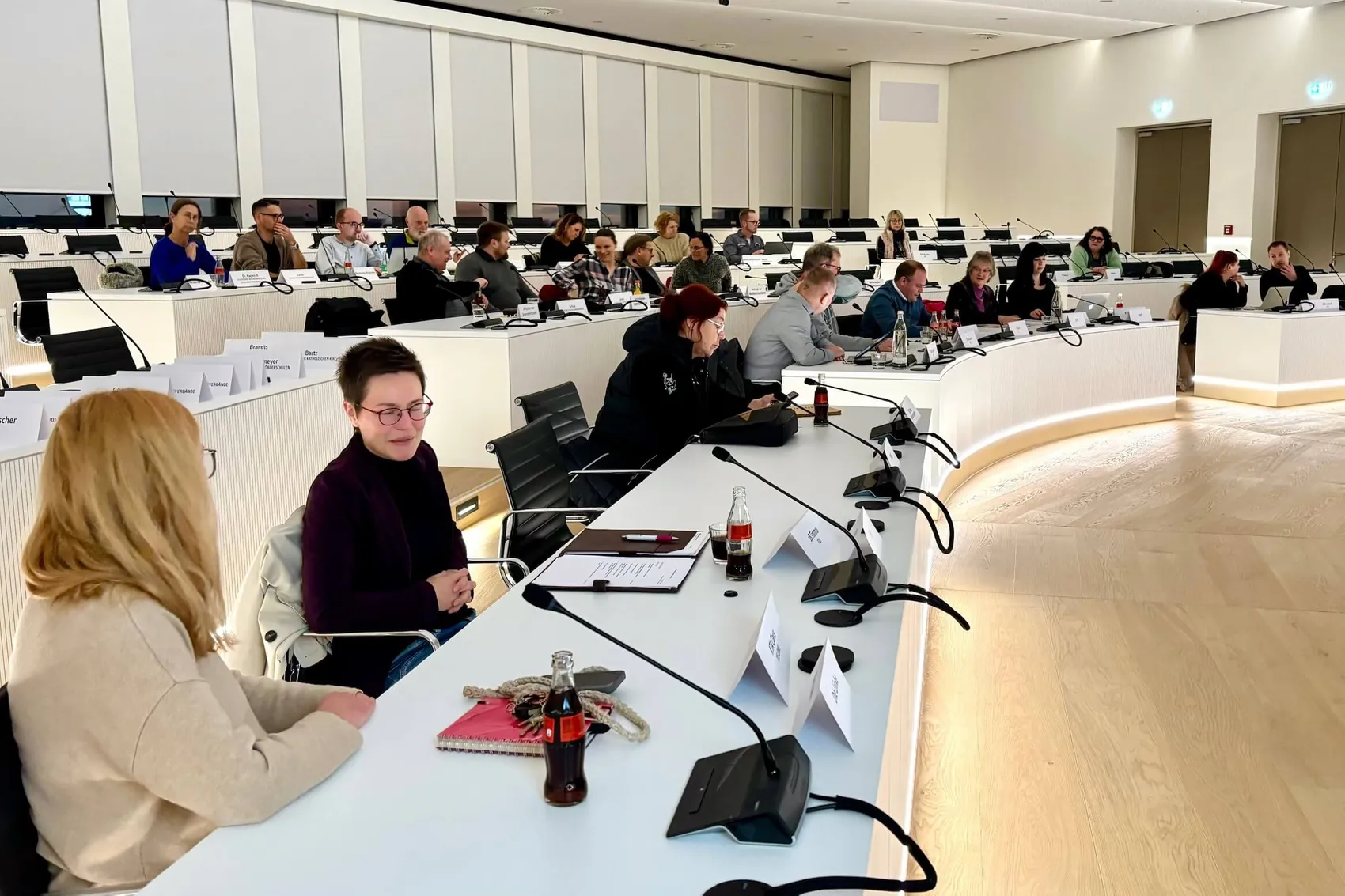 Versammlung im hellen Besprechungsraum mit langem Tisch und vielen Teilnehmern. Im Vordergrund sitzen zwei Frauen, die sich unterhalten, während auf dem Tisch Notizen, Getränke und Namensschilder liegen.