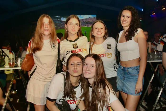 Fünf junge Frauen in weißen Trikots der deutschen Fußballnationalmannschaft posieren eng zusammen vor einer großen Leinwand, auf der ein Fußballspiel läuft. Im Hintergrund sind weitere Menschen und eine festliche Atmosphäre zu erkennen.