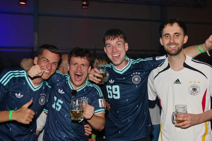 Vier junge Männer mit Biergläsern im Arm, gekleidet in Trikots des Deutschen Fußball-Bundes, lächeln in die Kamera. Im Hintergrund sind weitere feiernde Menschen zu erkennen.