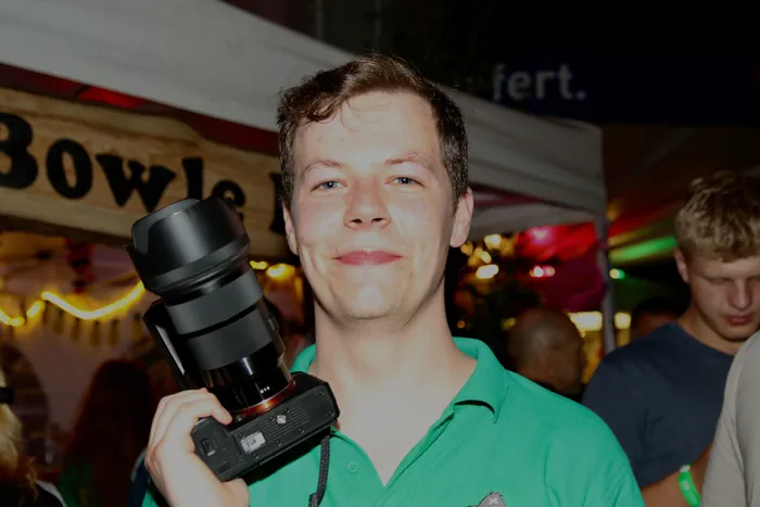 Ein junger Mann mit braunen Haaren hält eine professionelle Kamera vor sich. Im Hintergrund sind Menschen und ein beleuchteter Stand mit der Aufschrift 