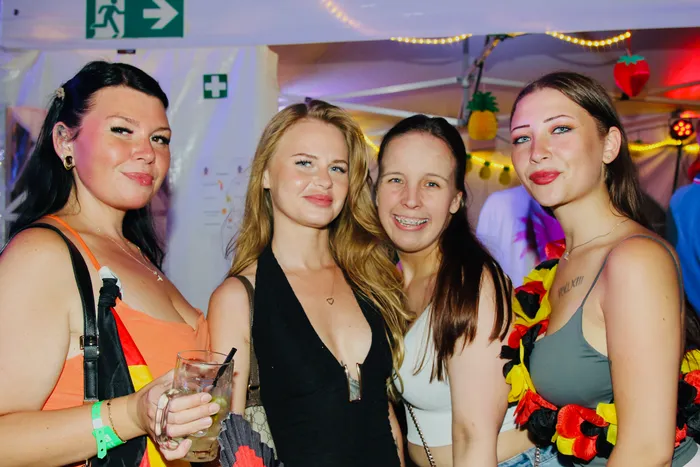 Vier junge Frauen stehen eng zusammen und lächeln in die Kamera. Sie tragen sommerliche Kleidung und Accessoires in den Farben der deutschen Flagge. Im Hintergrund sind Lichterketten und Dekorationen zu sehen.