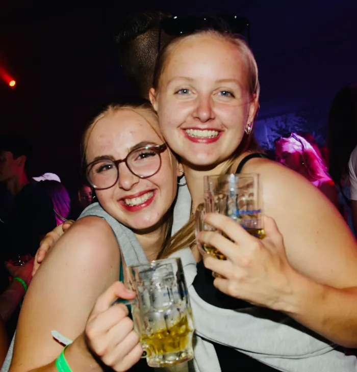 Zwei junge Frauen umarmen sich lachend und halten jeweils ein Bierglas in der Hand. Im Hintergrund sind weitere Menschen auf dem Scheunenfest der Landjugend Breckerfeld zu sehen.