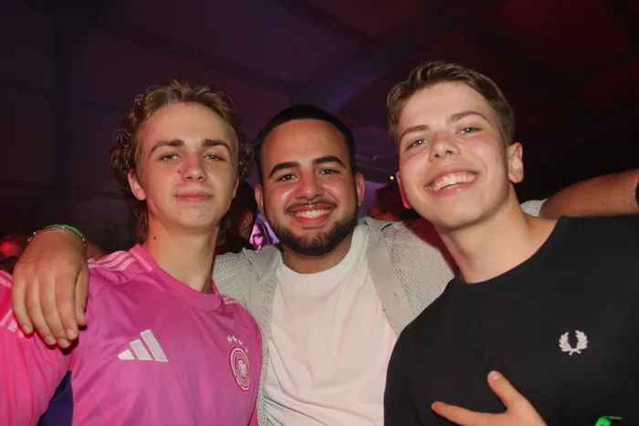 Drei junge Männer lächeln in die Kamera. Links trägt einer ein pinkes Fußballtrikot, in der Mitte ein weißes T-Shirt mit einem Cardigan darüber und rechts ein schwarzes Shirt mit einem Logo.