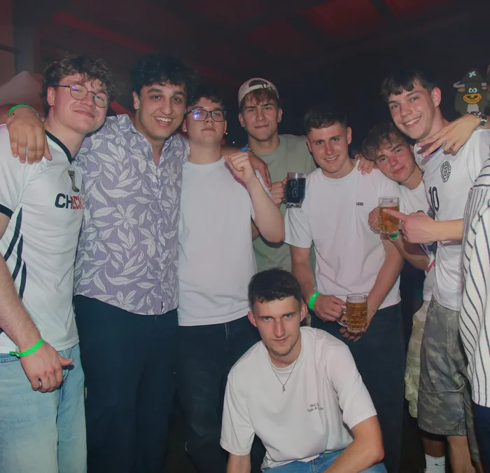 Eine Gruppe junger Männer steht eng zusammen und lächelt in die Kamera. Einige halten Biergläser in der Hand, andere haben ihre Arme um die Schultern der anderen gelegt. Im Hintergrund ist ein gelbes Stofftier zu sehen.