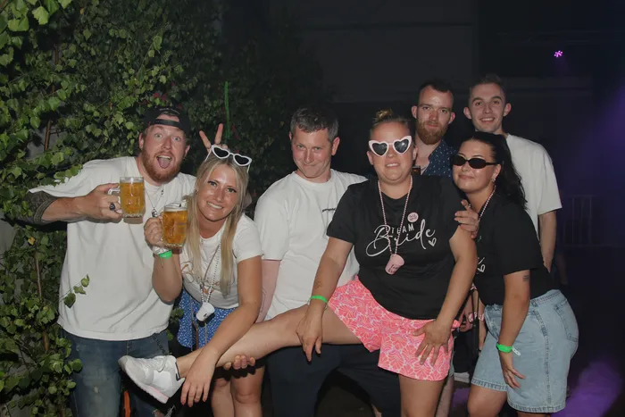 Eine Gruppe junger Erwachsener posiert lächelnd für ein Foto. Die Frau in der Mitte trägt ein T-Shirt mit der Aufschrift „Team Bride“ und eine rosa Shorts. Einige halten Bierkrüge in der Hand. Im Hintergrund ist üppiges Grün und ein beleuchtetes Gebäude zu sehen.
