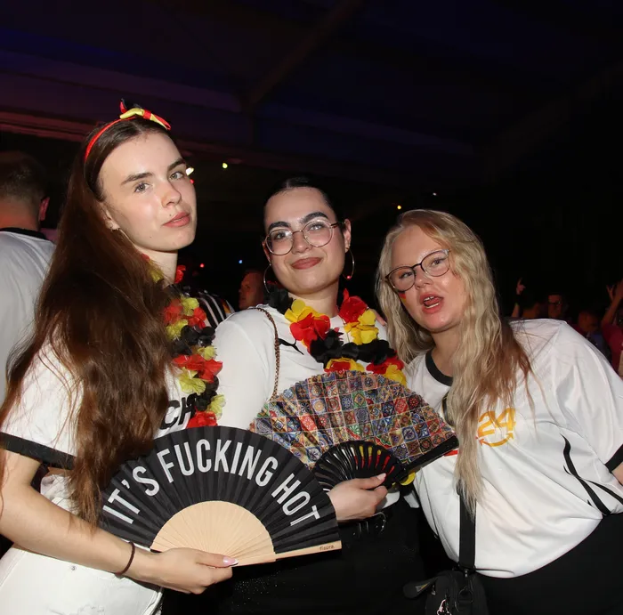 Drei junge Frauen mit Girlanden und Accessoires in den deutschen Nationalfarben posieren eng zusammen auf dem Scheunenfest der Landjugend Breckerfeld. Eine von ihnen hält einen Fächer mit der Aufschrift 