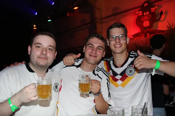 Drei junge Männer in Deutschlandtrikots stehen eng zusammen und halten jeweils ein Bierglas hoch. Im Hintergrund ist ein belebter Raum mit weiteren Personen und Dekorationen zu sehen.