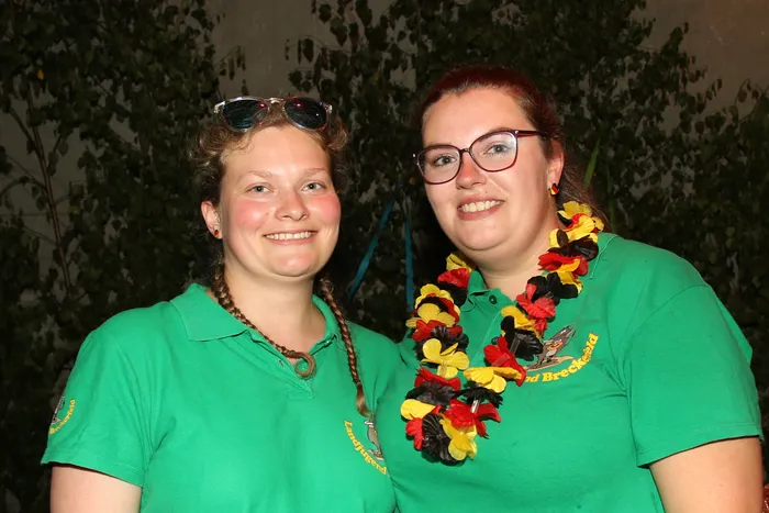 Zwei lächelnde junge Frauen mit grünen Poloshirts und deutschen Girlanden um den Hals. Im Hintergrund verschwommene Bäume und Lichter, passend zum Scheunenfest der Landjugend Breckerfeld.