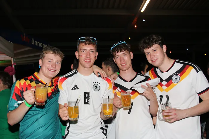 Vier junge Männer lächeln in die Kamera und halten Biergläser in der Hand. Sie tragen Trikots der deutschen Nationalmannschaft und Fanschminke in den Landesfarben. Im Hintergrund ist ein Festzelt mit der Aufschrift „Linnepe“ zu sehen.