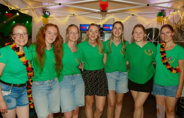 Sieben junge Frauen stehen lächelnd in einem Zelt. Sie tragen grüne Poloshirts mit einem Wappen und Jeansshorts, einige haben Girlanden um den Hals. Im Hintergrund sind Kühlschränke und Dekorationen zu sehen.
