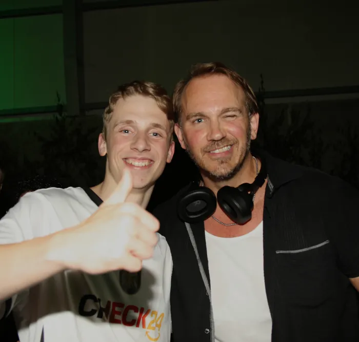 Zwei Männer posieren lächelnd für ein Foto. Der jüngere Mann zeigt den Daumen nach oben, während der ältere Mann Kopfhörer um den Hals trägt. Im Hintergrund ist ein grüner Bereich und ein dunkler Raum zu sehen.