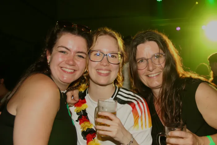 Drei lächelnde Frauen posieren eng zusammen vor einer verschwommenen Menschenmenge. Die Frau in der Mitte trägt ein Trikot der deutschen Nationalmannschaft und hält ein Bierglas, während die anderen beiden Frauen ebenfalls Getränke halten und eine Deutschlandkette tragen.