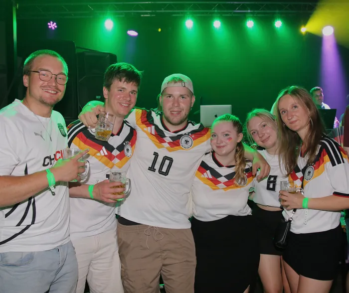 Eine Gruppe junger Erwachsener steht eng zusammen und lächelt in die Kamera. Viele tragen Trikots der deutschen Fußballnationalmannschaft und halten Biergläser in der Hand, während im Hintergrund eine Bühne mit grüner Beleuchtung zu sehen ist.