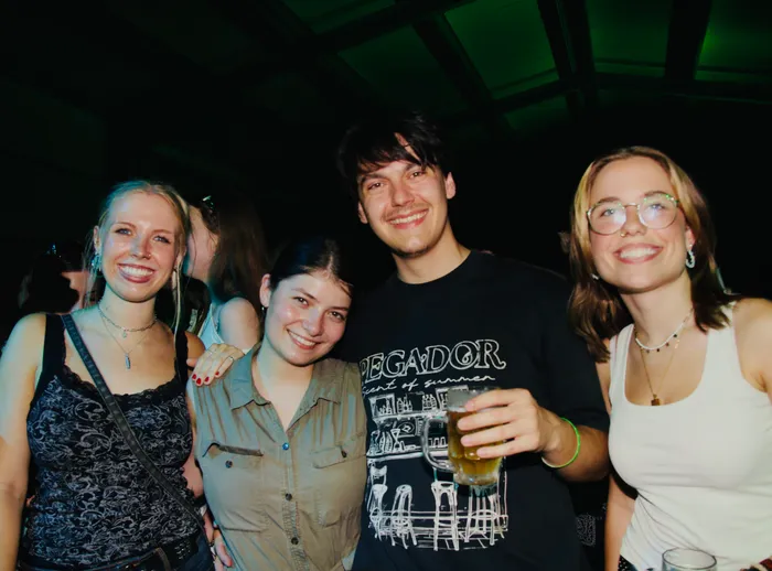 Vier junge Erwachsene lächeln in die Kamera. Der Mann in der Mitte hält ein Bierglas in der Hand, sein T-Shirt zeigt eine Skyline. Im Hintergrund ist ein dunkler Raum mit grünem Licht zu sehen.