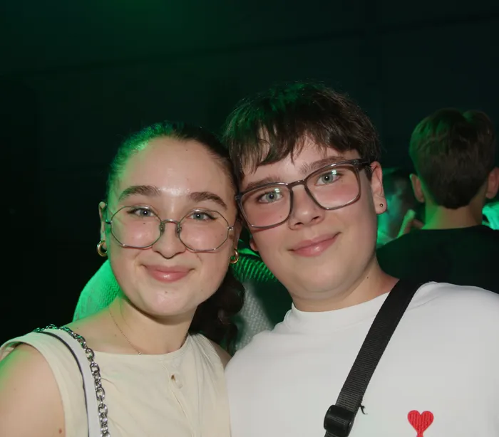 Zwei junge Menschen mit Brille schauen in die Kamera. Im Hintergrund ist ein grünes Licht und weitere Personen auf einem Scheunenfest zu erkennen.