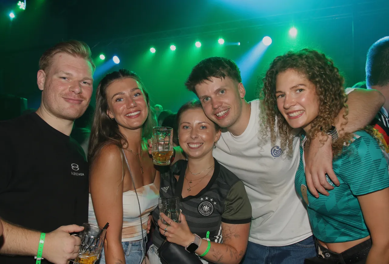 Fünf junge Erwachsene lächeln in die Kamera, umgeben von Menschen in einem dunklen Raum mit Bühnenbeleuchtung. Eine Frau trägt das Trikot der deutschen Fußballnationalmannschaft.