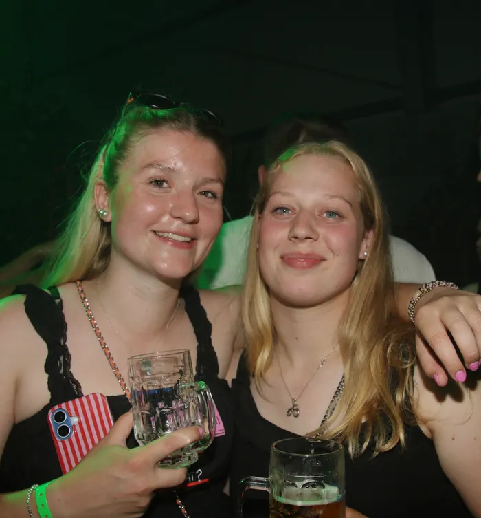 Zwei junge Frauen mit Bierkrügen posieren eng umschlungen auf dem Scheunenfest der Landjugend Breckerfeld. Im Hintergrund sind weitere Menschen und eine grünlich beleuchtete Umgebung zu erkennen.