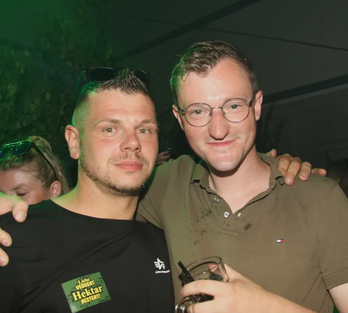 Zwei junge Männer posieren eng umschlungen vor der Kamera. Der Mann links trägt ein schwarzes T-Shirt mit Aufschrift und der Mann rechts ein olivgrünes Poloshirt. Im Hintergrund sind weitere Personen und grüne Beleuchtung zu sehen.