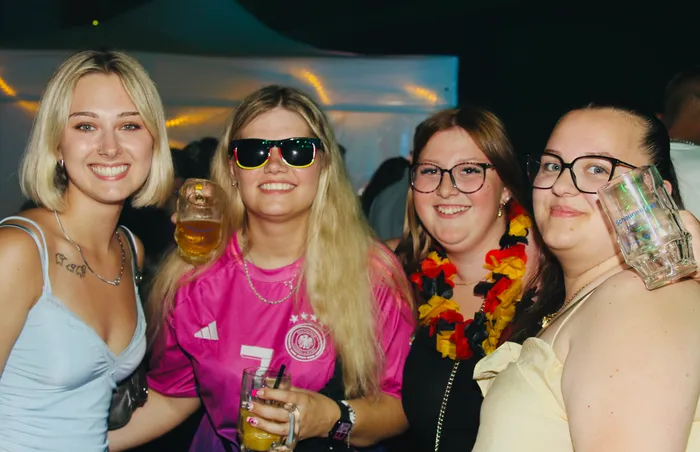 Vier junge Frauen stehen eng zusammen und lächeln in die Kamera. Zwei von ihnen tragen Deutschland-Trikots, eine weitere eine Deutschland-Fahne um den Hals, und alle halten Bier oder Mixgetränke in der Hand. Im Hintergrund ist ein festlich beleuchteter Zeltraum zu erkennen.