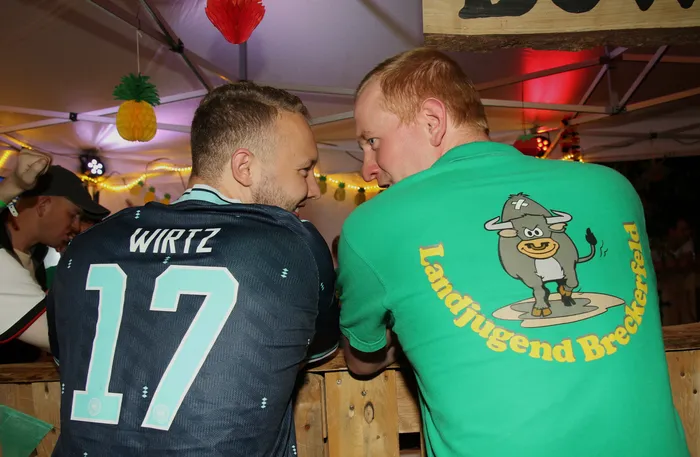 Zwei Männer stehen sich auf einem Scheunenfest gegenüber. Der Mann links trägt ein Fußballtrikot mit der Aufschrift 