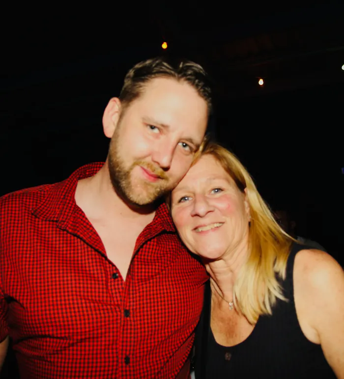 Ein Mann mit rotem kariertem Hemd und eine ältere Frau mit blonden Haaren lächeln in die Kamera und posieren eng aneinander. Im Hintergrund ist ein dunkler Raum mit wenigen Lichtpunkten zu sehen.