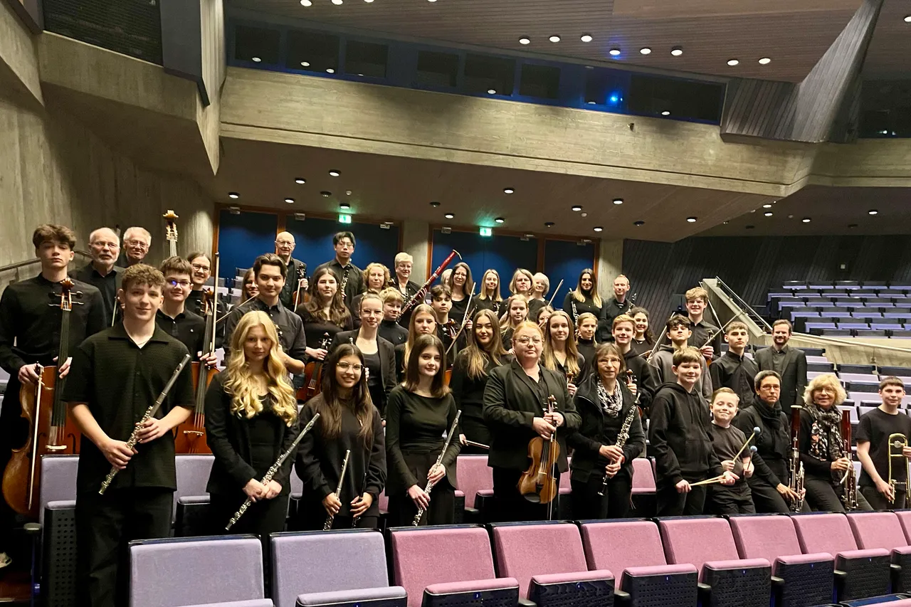 Große Gruppe junger Musiker mit verschiedenen Instrumenten posiert auf den Sitzen eines Konzerthauses.