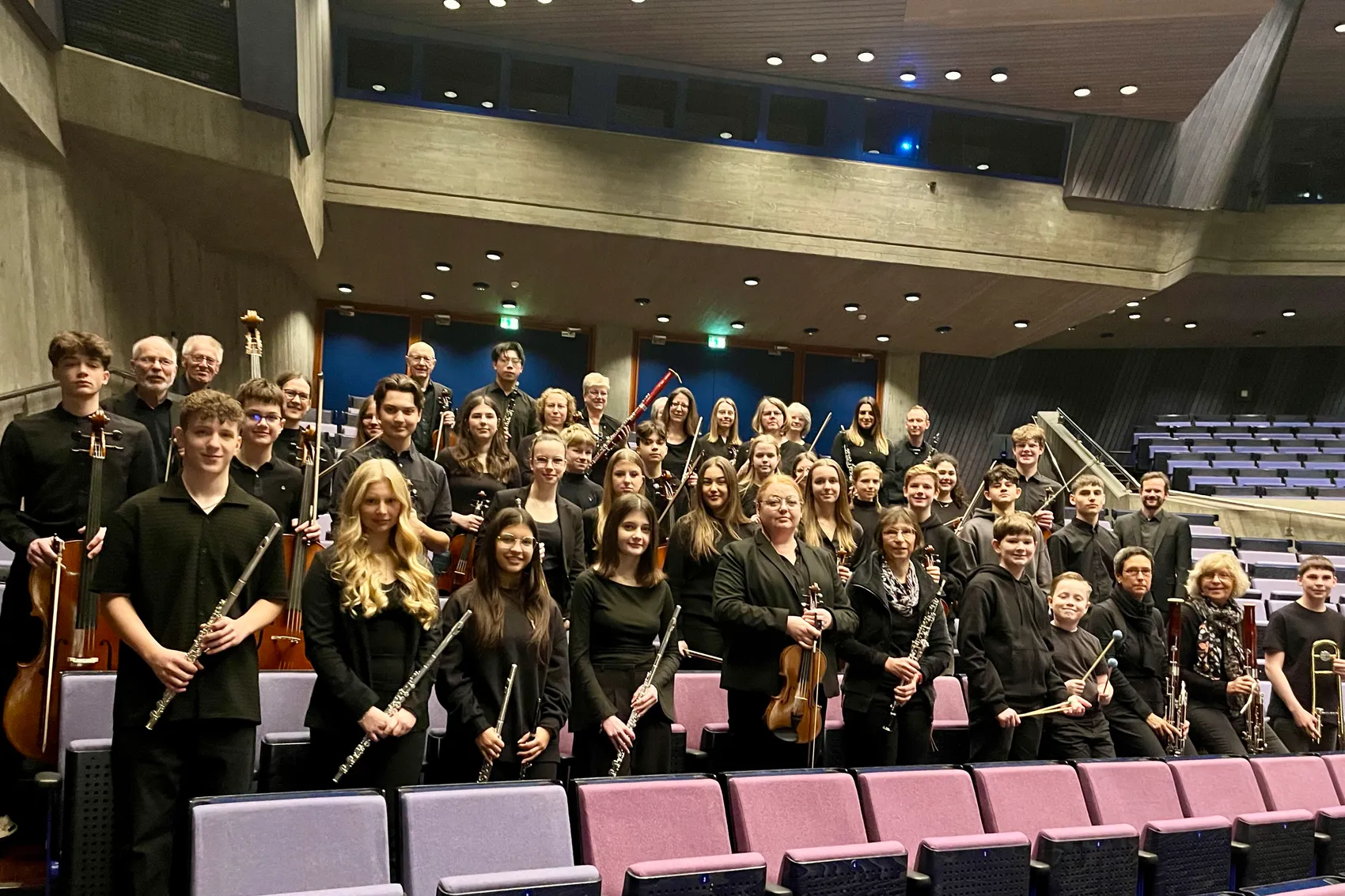 Große Gruppe junger Musiker mit verschiedenen Instrumenten posiert auf den Sitzen eines Konzerthauses.