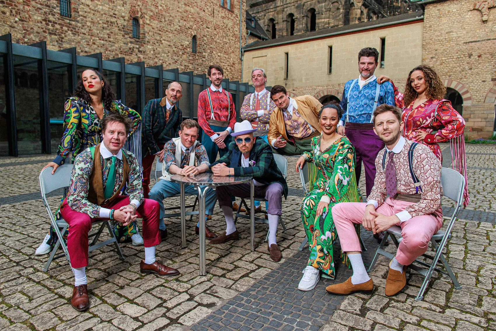 Die Band Jan Delay & Disko Nr. 1 posiert in farbenfrohen, gemusterten Outfits in einem gepflasterten Innenhof vor alten Backsteingebäuden.