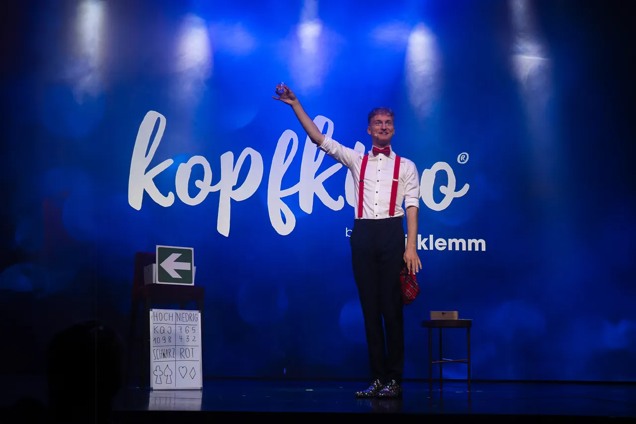 Jannis Klemm auf der Bühne mit erhobener Hand vor einem blauen Hintergrund mit dem Schriftzug „kopfkino“. Neben ihm ein Podest mit Karten und ein Mikrofon.