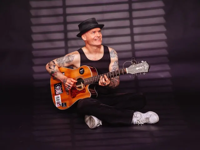 Tobias Janssen mit schwarzem Hut und Tätowierungen an den Armen sitzt auf dem Boden und spielt eine akustische Gitarre.