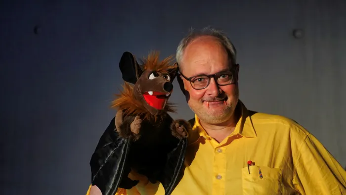 Joachim Hecker lächelt und hält eine braune Fledermaus-Handpuppe vor einem dunklen Hintergrund.