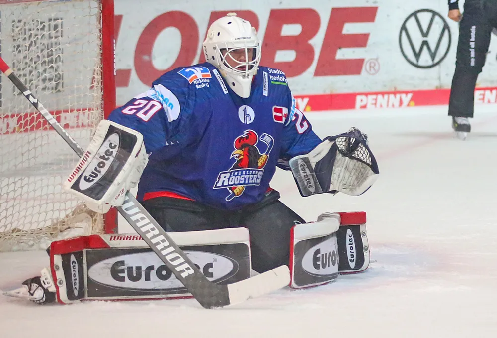 Eishockeytorwart Jonas Neffin in voller Ausrüstung, während er im Tor der Iserlohn Roosters steht und den Schläger hält. Im Hintergrund ist Werbebeschilderung zu sehen.
