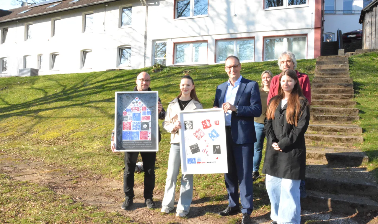 Fünf Personen und zwei Jugendliche halten Linoldrucke vor einem hellen Gebäude auf einer Anhöhe. Die Drucke zeigen abstrakte Muster und Porträts.