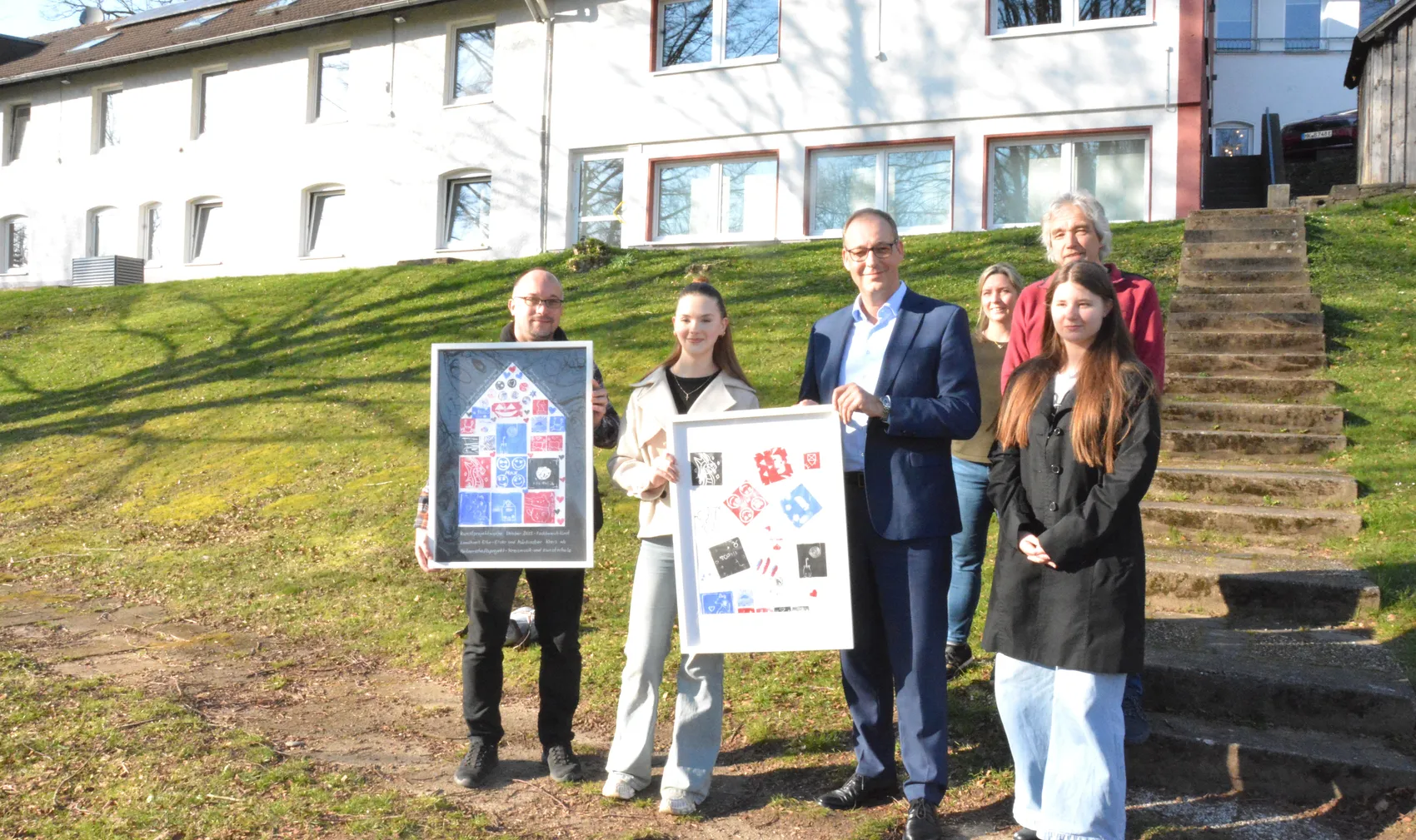 Fünf Personen und zwei Jugendliche halten Linoldrucke vor einem hellen Gebäude auf einer Anhöhe. Die Drucke zeigen abstrakte Muster und Porträts.