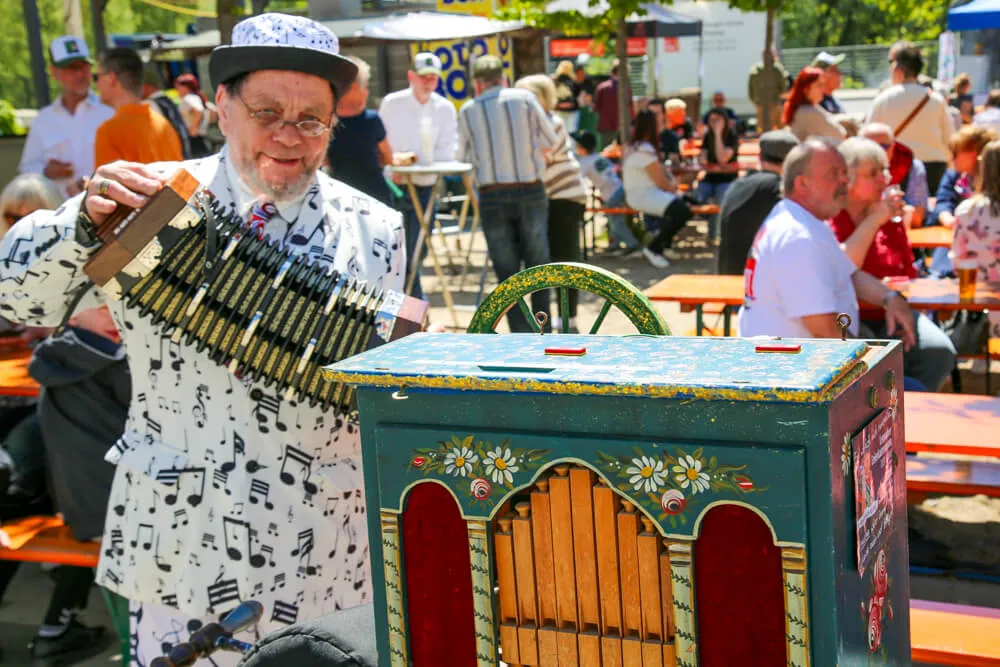 Ein älterer Mann mit Hut spielt auf einem verzierten Handorgel vor Menschen an Biertischen im Freien.