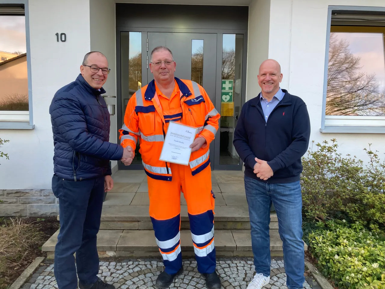 Drei Männer stehen vor einem hellen Gebäude und lächeln in die Kamera. Der Mann in der Mitte hält eine Urkunde und trägt einen orangefarbenen Arbeitsanzug, während die Männer links und rechts ihn mit einem Händedruck und freundlichem Blick beglückwünschen.