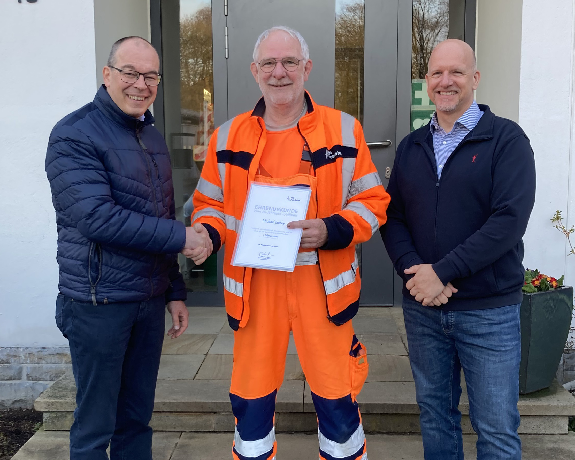 Drei Männer im Freien: Marco Gräb und Jens Kaminski überreichen Michael Jacoby eine Urkunde für 25 Jahre Dienstjubiläum.