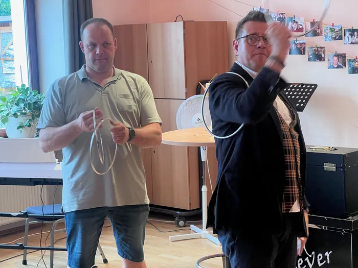 Ein Mann in einem grünen Poloshirt klatscht in die Hände, während ein Mann in einem schwarzen Anzug mit einem Seil jongliert. Im Hintergrund steht ein Ventilator und eine Pinnwand mit Fotos.