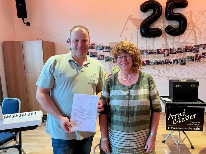 Ein Mann und eine Frau stehen nebeneinander und halten ein Dokument. Im Hintergrund befindet sich eine goldene „25“-Folie und ein Keyboard.