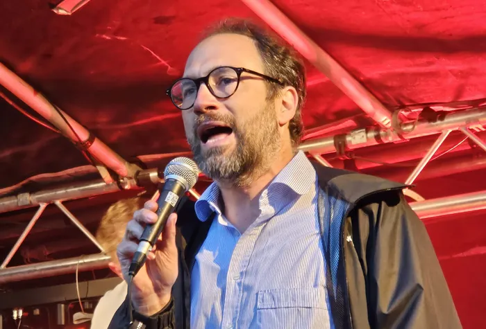 Mann mit Brille singt in ein Mikrofon auf einer Bühne vor rotem Hintergrund.