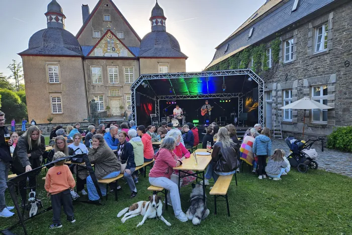 Open-Air-Konzert vor einem historischen Schloss mit Zuschauern an Biertischen und stehenden Besuchern, zwei Hunde im Vordergrund.