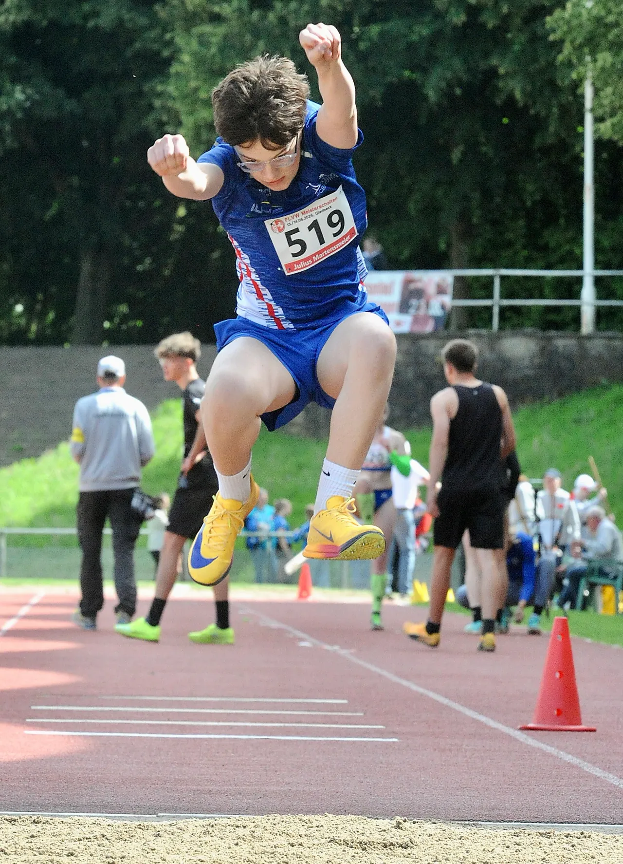 Ein Jugendlicher in Sportkleidung springt beim Dreisprung in der Luft. Er trägt die Startnummer 519 und gelbe Sportschuhe. Im Hintergrund sind weitere Sportler und Zuschauer zu sehen.