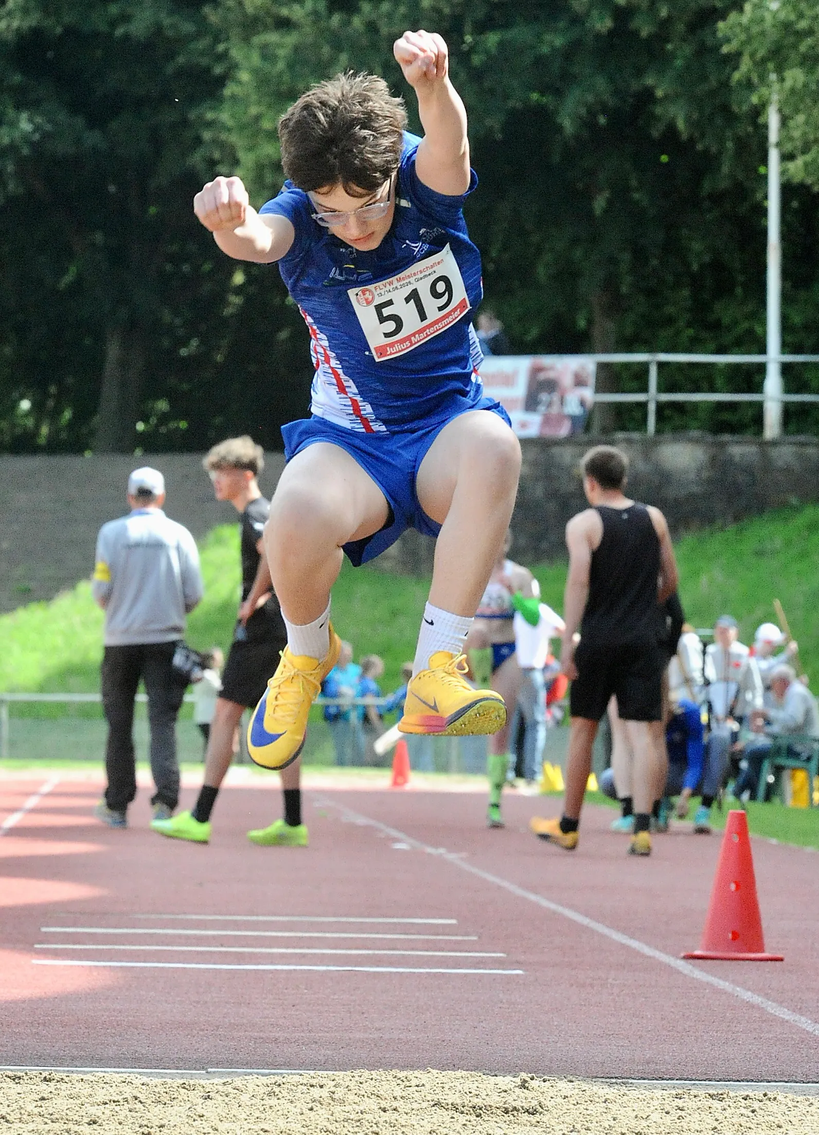 Ein Jugendlicher in Sportkleidung springt beim Dreisprung in der Luft. Er trägt die Startnummer 519 und gelbe Sportschuhe. Im Hintergrund sind weitere Sportler und Zuschauer zu sehen.